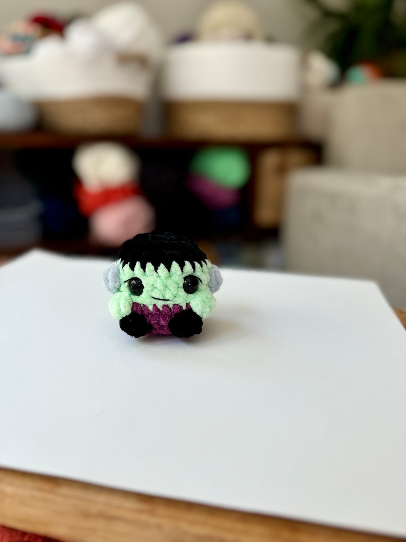 May include: A small green crocheted monster with black hair, purple clothing, and big black eyes. The monster is sitting on a white surface.