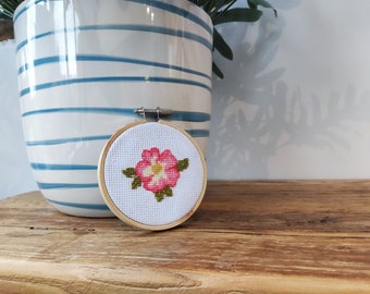 May include: A small, round, wooden embroidery hoop with a white fabric background and a pink flower with green leaves embroidered in the center.