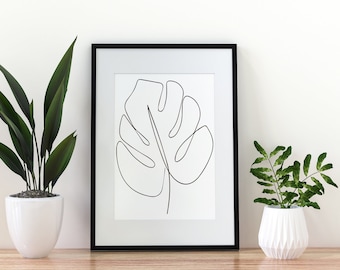 May include: A black framed print of a single line drawing of a monstera leaf, hanging on a white wall between two potted plants. The plants are in white ceramic pots.