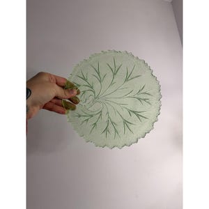 Puede incluir: Un plato redondo de vidrio verde claro con una superficie texturizada y un diseño decorativo de hojas. El plato tiene un borde festoneado y está sostenido por una mano. El diseño de hojas está grabado en el vidrio.