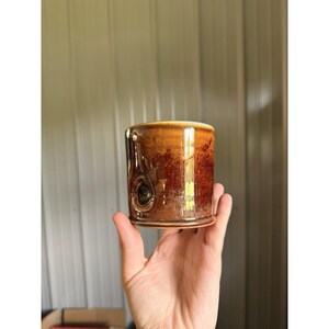 May include: A cylindrical ceramic mug featuring a mottled brown and tan glaze with a smooth, glossy finish. The exterior surface displays a distinct, dark circular indentation resembling a knot, with a lighter rim at the top and base.