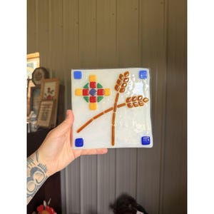 May include: A square fused glass tile with a white textured base. The design features two crossed wheat stalks in brown, a geometric star pattern made of small red, yellow, green, and purple squares, and four blue glass squares in the corners.