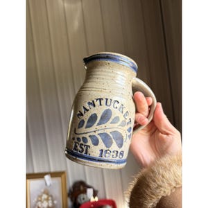 May include: A beige ceramic pitcher with a blue rim and handle. The pitcher is decorated with the word "Nantucket" in blue, along with a leaf design and the text "EST. 1638". The pitcher is held by a person.