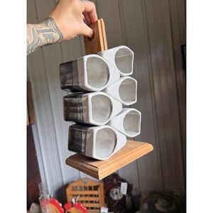 May include: A set of six white and dark gray ceramic mugs stacked in two vertical columns on a light brown wooden stand. The mugs have a smooth, glossy finish with a distinct color-blocked design, featuring white interiors and dark gray exteriors.
