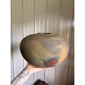May include: A round, low-profile ceramic vase with a matte, multi-colored finish featuring blended tones of muted brown, deep orange, and hints of dark blue. The vessel has a wide, bulbous body that tapers to a small, circular opening at the top.