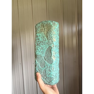 Puede incluir: Un bloque rectangular con apariencia de piedra y un acabado texturizado y desgastado en tonos turquesa y bronce. La superficie presenta tres diseños de mariposas en relieve de distintos tamaños dispuestos verticalmente en la cara frontal.