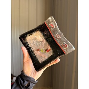 May include: A square fused glass dish featuring a black center panel with a textured beige fabric inlay, a silver spiral design, and small red and green accents. The border is a speckled silver glass with a thin red line and three small square insets.