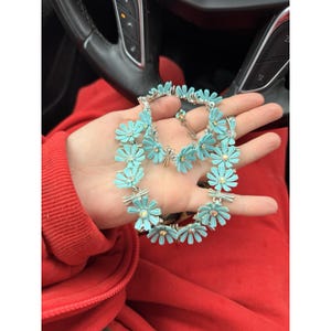 Könnte beinhalten: Ein türkisfarbenes Halsketten- und Armband-Set mit Gänseblümchen-Blüten. Der Schmuck hat silberfarbenes Metall und kleine, funkelnde Akzente in der Mitte jeder Blüte. Das Set wird auf einer Hand vor rotem Hintergrund präsentiert.