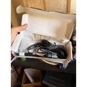 Puede incluir: Una caja de cartón blanca que contiene un cable eléctrico negro enrollado con un arnés de cables multicolor y un conector de plástico negro. Dentro de la caja hay una pequeña tarjeta de instrucciones con pasos numerados y texto de instalación.