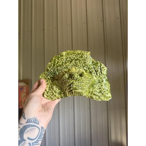 May include: A semi-circular ceramic wall plaque in a vibrant lime green glaze. The surface features a highly textured, organic relief pattern with a central raised element and a small circular hole at the top for hanging purposes.