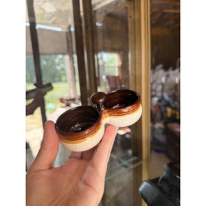 May include: A small, dual-compartment ceramic dish featuring a cream-colored exterior and a dark brown glazed interior. The two circular bowls are connected by a central handle, creating a functional piece with a smooth, glossy finish.