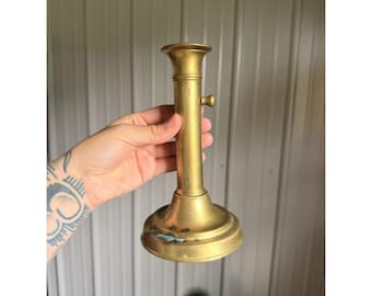 May include: A brass candlestick holder featuring a cylindrical stem, a flared top rim, and a wide circular base. The metal surface displays a smooth finish with visible signs of oxidation and patina near the bottom edge of the base.