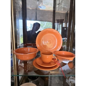 May include: A set of orange ceramic dinnerware including a large dinner plate, a smaller salad plate, a bowl, and a cup with a matching saucer. The pieces feature a smooth, glossy finish and a solid, vibrant orange color throughout the collection.