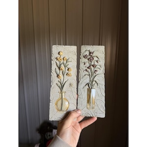 Peut inclure: Deux plaques murales rectangulaires avec une finition texturée gris clair rappelant la pierre. La plaque de gauche présente un relief de fleurs jaunes dans un vase rond en verre. Celle de droite montre des fleurs violettes dans un vase rectangulaire.