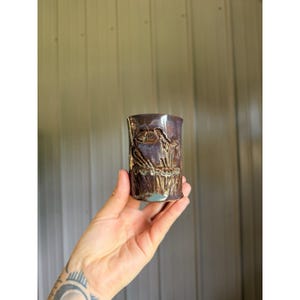 May include: A cylindrical ceramic cup with a glossy, deep purple glaze. The exterior features a textured, relief-style design depicting a bird perched among tall grasses, with subtle brown and metallic highlights accentuating the carved details.