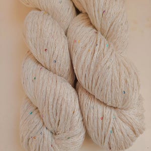 Pu&ograve; includere: Matassa di filato bianco sporco con paillettes colorate. Il filato &egrave; attorcigliato strettamente, mostrando la trama delle fibre. Le paillettes, piccole, sono sparse, aggiungendo un tocco di brillantezza. Adatto per progetti di maglia o uncinetto.