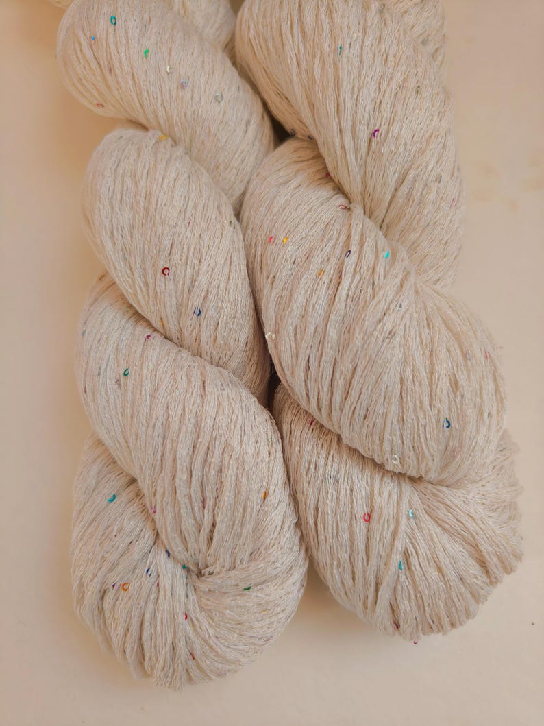 Pu&ograve; includere: Matassa di filato bianco sporco con paillettes colorate. Il filato &egrave; attorcigliato strettamente, mostrando la trama delle fibre. Le paillettes, piccole, sono sparse, aggiungendo un tocco di brillantezza. Adatto per progetti di maglia o uncinetto.
