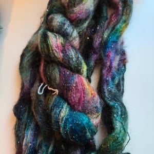 Peut inclure: Un écheveau de laine filée à la main, tressé et épais, présentant une palette de couleurs variées de vert forêt profond, bleu roi, magenta et gris anthracite. La fibre texturée et duveteuse est parsemée de petites paillettes argentées.