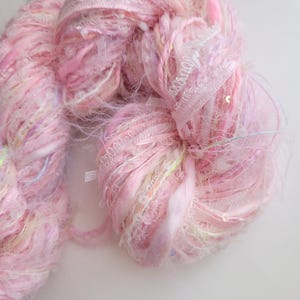 Peut inclure: Gros plan sur une pelote de fil rose avec des accents blancs, jaunes et bleus. Le fil a un aspect texturé et duveteux avec des rubans et des paillettes. Ce fil est adapté aux projets de tricot et de crochet.