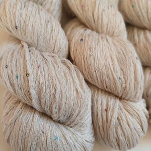 Pu&ograve; includere: Primo piano di matasse di filato beige con paillettes colorate. Il filato appare morbido e strutturato, ideale per progetti di maglieria o uncinetto. Le paillettes aggiungono un tocco di brillantezza al filato.