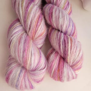 May include: Two skeins of hand-dyed yarn in shades of pink, purple, and white. The yarn has a soft, fluffy texture with subtle color variations. Ideal for knitting or crocheting projects.