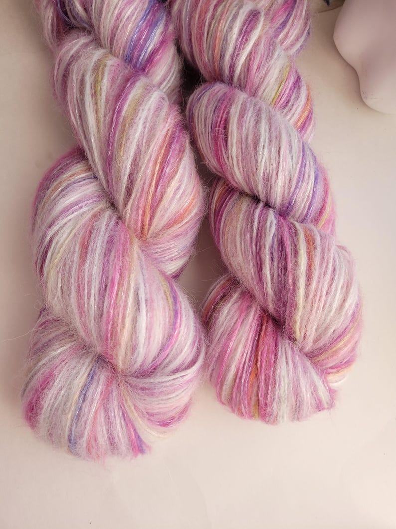 May include: Two skeins of hand-dyed yarn in shades of pink, purple, and white. The yarn has a soft, fluffy texture with subtle color variations. Ideal for knitting or crocheting projects.