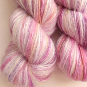 May include: Close-up of several skeins of yarn in shades of pink, white, and purple. The yarn has a soft, fluffy texture with a subtle shimmer. The colors blend together, creating a visually appealing and delicate appearance. Ideal for knitting or crochet projects.