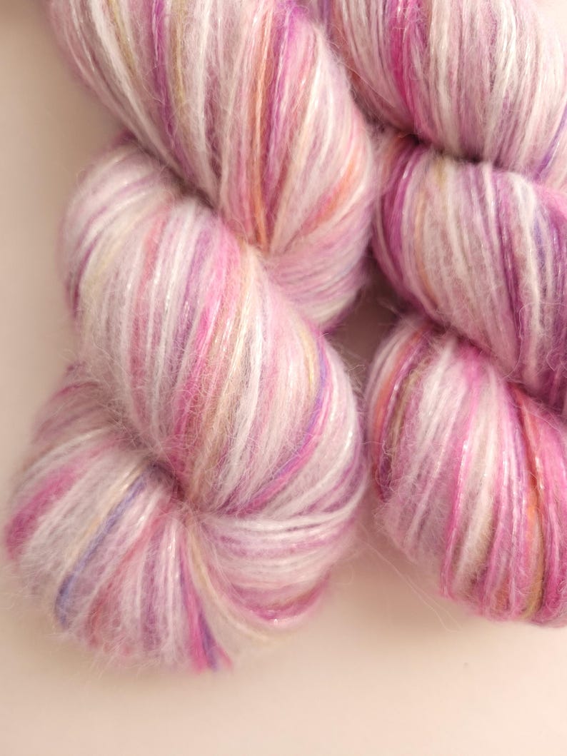 May include: Close-up of several skeins of yarn in shades of pink, white, and purple. The yarn has a soft, fluffy texture with a subtle shimmer. The colors blend together, creating a visually appealing and delicate appearance. Ideal for knitting or crochet projects.