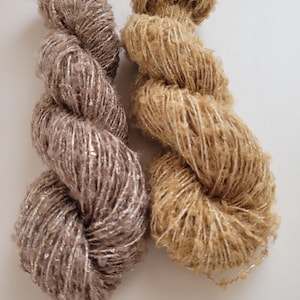Peut inclure: Deux écheveaux de fil, l'un brun clair et l'autre beige clair. Les deux fils ont un aspect texturé et duveteux avec des brins dorés métalliques tissés, créant un effet scintillant. Le fil est probablement destiné aux projets de tricot ou de crochet.