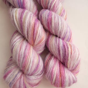 May include: Two skeins of hand-dyed yarn in shades of pink, purple, and white. The yarn has a soft, fluffy texture and a subtle sheen. The colors blend together in a variegated pattern, creating a visually appealing effect. Ideal for knitting or crocheting projects.