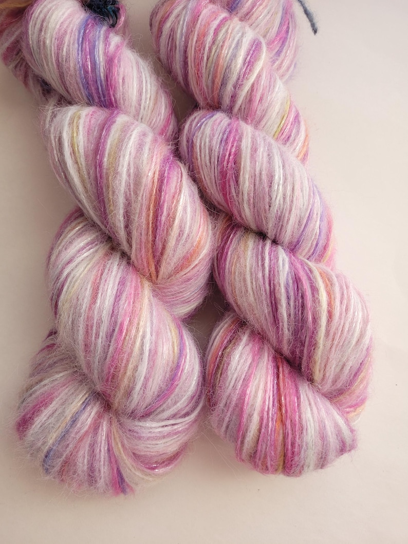 May include: Two skeins of hand-dyed yarn in shades of pink, purple, and white. The yarn has a soft, fluffy texture and a subtle sheen. The colors blend together in a variegated pattern, creating a visually appealing effect. Ideal for knitting or crocheting projects.