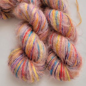 May include: Close-up of several skeins of yarn. The yarn is a soft, fluffy texture with a light pink base color. Strands of vibrant colors, including red, yellow, blue, and purple, are interwoven throughout the yarn, creating a colorful effect.