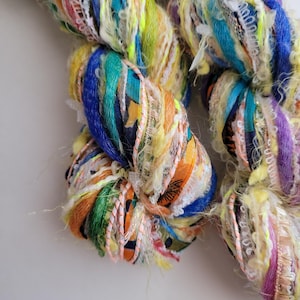 May include: Close-up of a colourful yarn skein. The yarn features a mix of vibrant colours including blue, yellow, orange, green, and purple. The yarn has a textured appearance with various ribbons and threads.