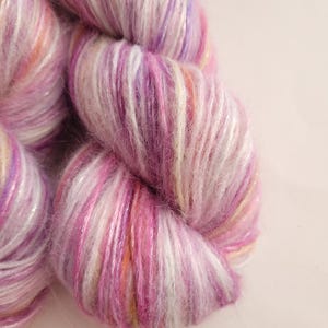 May include: Close-up of a skein of yarn in shades of pink, purple, and white. The yarn has a soft, textured appearance with subtle hints of orange and yellow. Ideal for knitting or crochet projects.