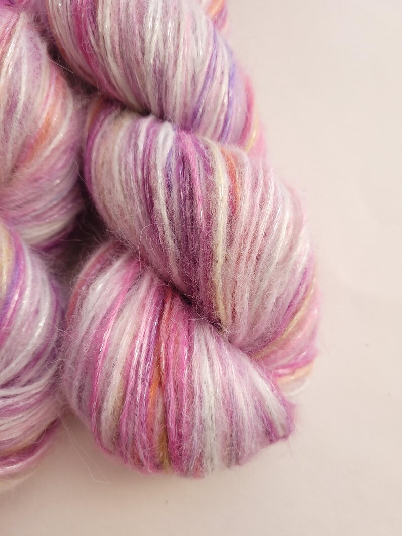 May include: Close-up of a skein of yarn in shades of pink, purple, and white. The yarn has a soft, textured appearance with subtle hints of orange and yellow. Ideal for knitting or crochet projects.