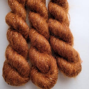 May include: Three skeins of copper-colored yarn with a metallic sheen. The yarn is tightly twisted, creating a textured appearance. The close-up shot highlights the yarn's color and the subtle sparkle of the metallic threads, perfect for knitting or crochet projects.