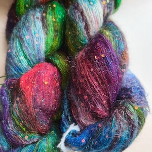 May include: Two skeins of fuzzy, multi-colored yarn featuring a blend of green, blue, purple, and red fibers. The yarn is interwoven with shimmering metallic threads and small, flat, flower-shaped sequins scattered throughout the textured strands.