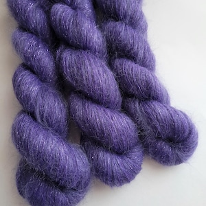 May include: Close-up of three skeins of purple yarn with a shimmering, metallic thread woven throughout. The yarn is thick and fluffy, with a soft, textured appearance. The color is a deep, rich purple, with silver highlights.