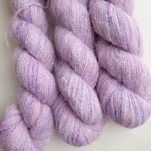 May include: Close-up of three skeins of lavender yarn with a soft, textured appearance. The yarn has a subtle sheen with silver and pink threads interwoven throughout. The twisted strands create a visually appealing, fluffy texture, ideal for knitting or crochet projects.