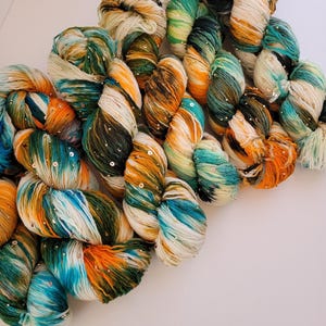 Peut inclure: Gros plan sur plusieurs écheveaux de fil aux couleurs variées : blanc, orange, turquoise et vert. Le fil est parsemé de petites paillettes argentées. Le fil est disposé en diagonale sur un fond blanc.