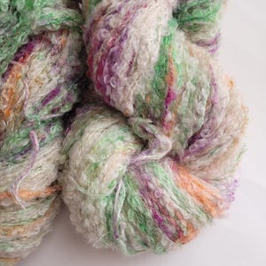May include: Close-up of two skeins of colorful yarn. The yarn is a mix of green, white, purple, and orange. The texture is thick and textured, with a slightly fuzzy appearance. The yarn appears to be for knitting or crocheting.