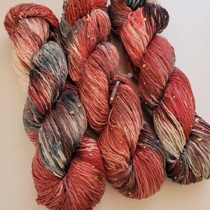 May include: Several skeins of yarn in shades of red, gray, and cream, speckled with iridescent sequins. The yarn is arranged in loose, twisted bundles, showcasing the color variations and texture. Ideal for knitting or crochet projects.
