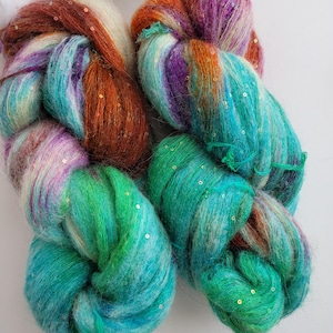 May include: Two skeins of yarn with a blend of colors including teal, green, purple, orange, and white. The yarn is embellished with gold sequins.