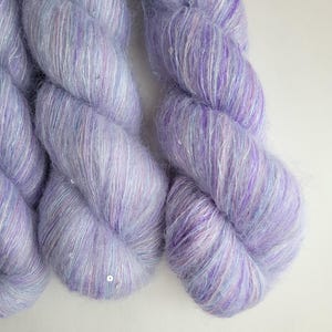May include: Close-up of three skeins of lavender yarn with a soft, fuzzy texture. The yarn has a subtle sheen and is interspersed with iridescent threads and small sequins. The yarn is ideal for knitting or crocheting projects.