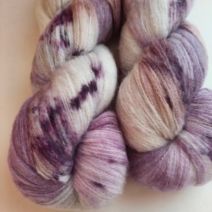 Peut inclure: Deux écheveaux de fil doux et duveteux présentant une palette de couleurs variées composée de crème, de rose clair et de violet profond. Les fibres ont une texture brossée semblable au mohair avec des transitions de couleurs visibles.