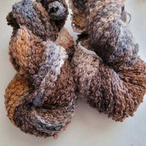 May include: Two skeins of textured yarn in shades of brown, tan, grey, and white. The yarn has a looped, curly texture, and is tightly wound. The skeins are on a white surface.