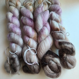 May include: Four skeins of hand-dyed yarn featuring a soft, fuzzy texture. The yarn displays a variegated color palette of cream, pale pink, lavender, and deep brown tones, with each skein twisted and secured with a white string tie.