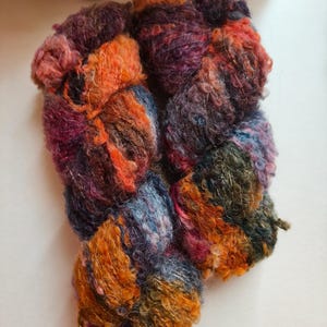 May include: A skein of textured yarn featuring a variegated colour palette of deep orange, dark purple, forest green, and muted blue. The yarn has a fuzzy, looped construction with subtle metallic threads woven throughout the fibres.