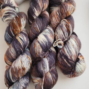 Op de afbeelding: Drie strengen handgeverfd garen in een mix van kleuren. Het garen heeft tinten bruin, paars, crème en een vleugje blauw. Het garen is tot een streng getwist, klaar voor brei- of haakprojecten.
