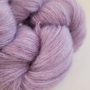 May include: Close-up of a skein of soft, lavender-coloured yarn. The yarn has a fluffy texture with subtle colour variations and a slight sheen. The fibres are tightly spun, creating a luxurious appearance, perfect for knitting or crochet projects.